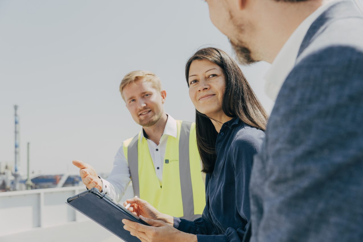 Maria Trinh, Roman Fritsches und Leon Brach mit Klemmbrett vor Industrieanlage